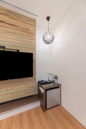 Una camera moderna con una TV a schermo piatto fissata a una parete in legno, un tavolino con una macchina del caffè e bottiglie d'acqua, e un frigorifero compatto.