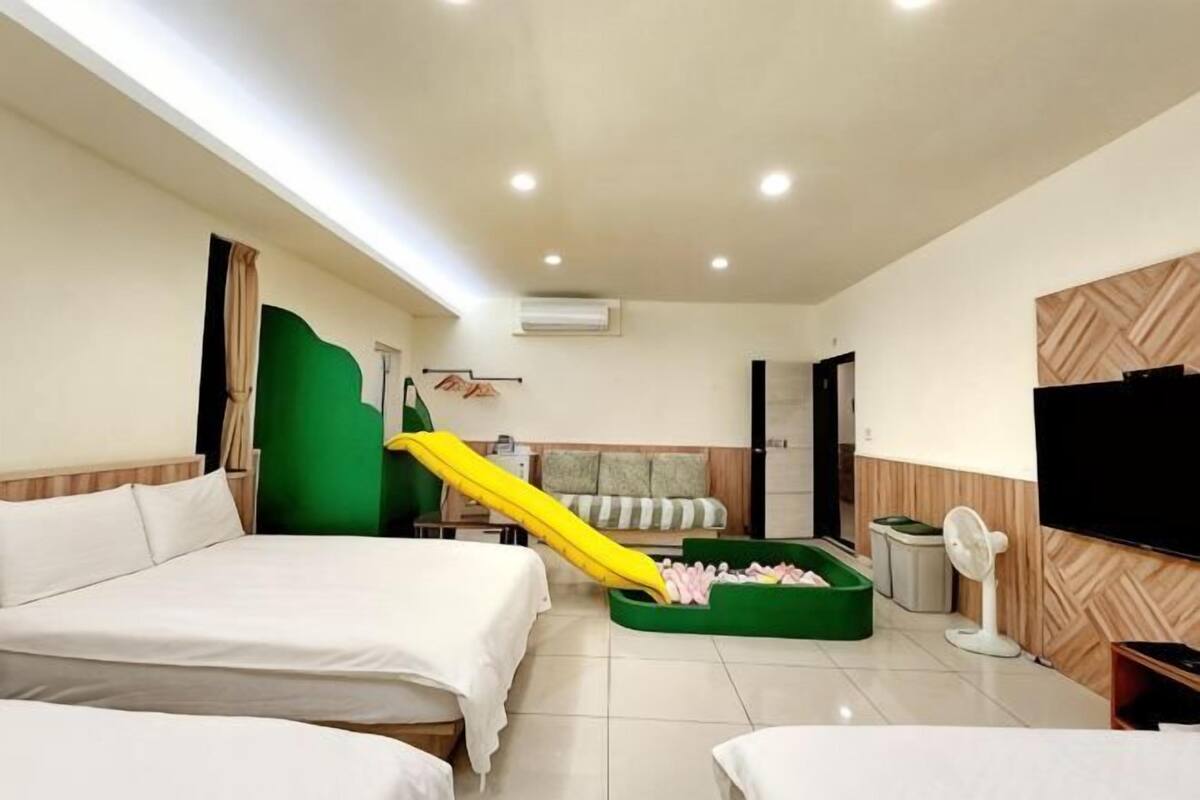 Hotellihuone, jossa on kaksi sänkyä, vihreä sohva, keltainen liukumäki, televisio ja tuuletin.