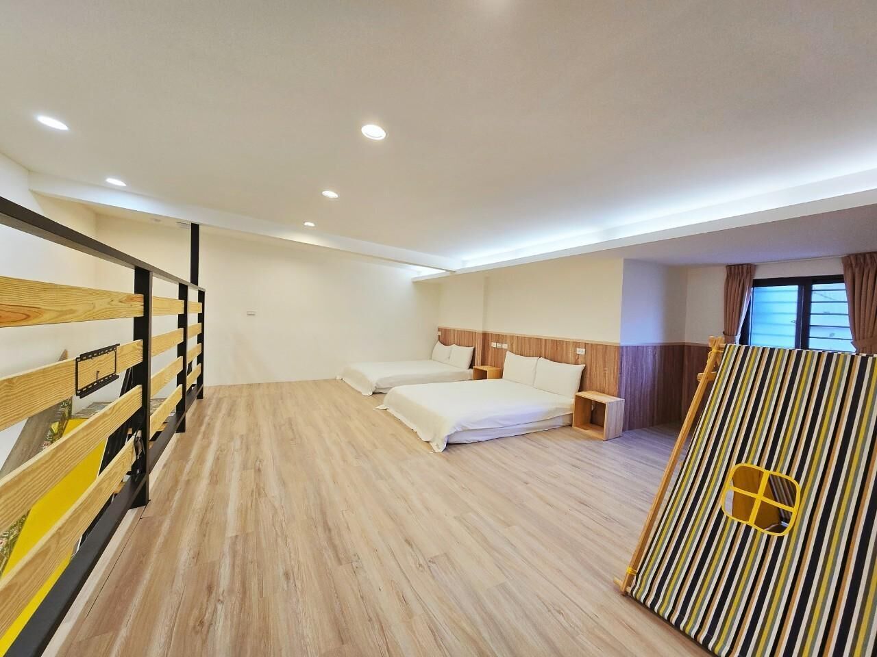 A hotel room with a wooden floor, two beds, a wooden bunk bed, a striped partition, and a window with curtains.