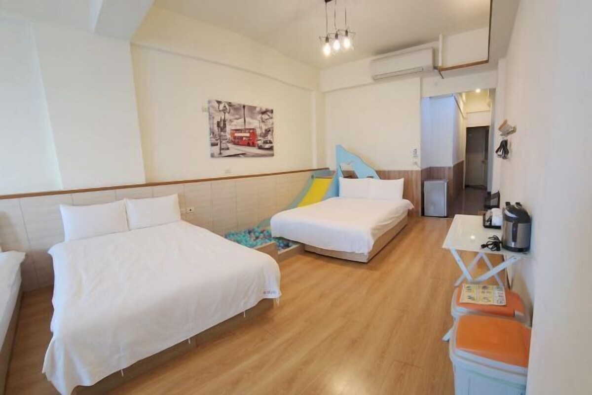 A hotel room with two beds, a small table, a chair, a heater, and a painting on the wall.
