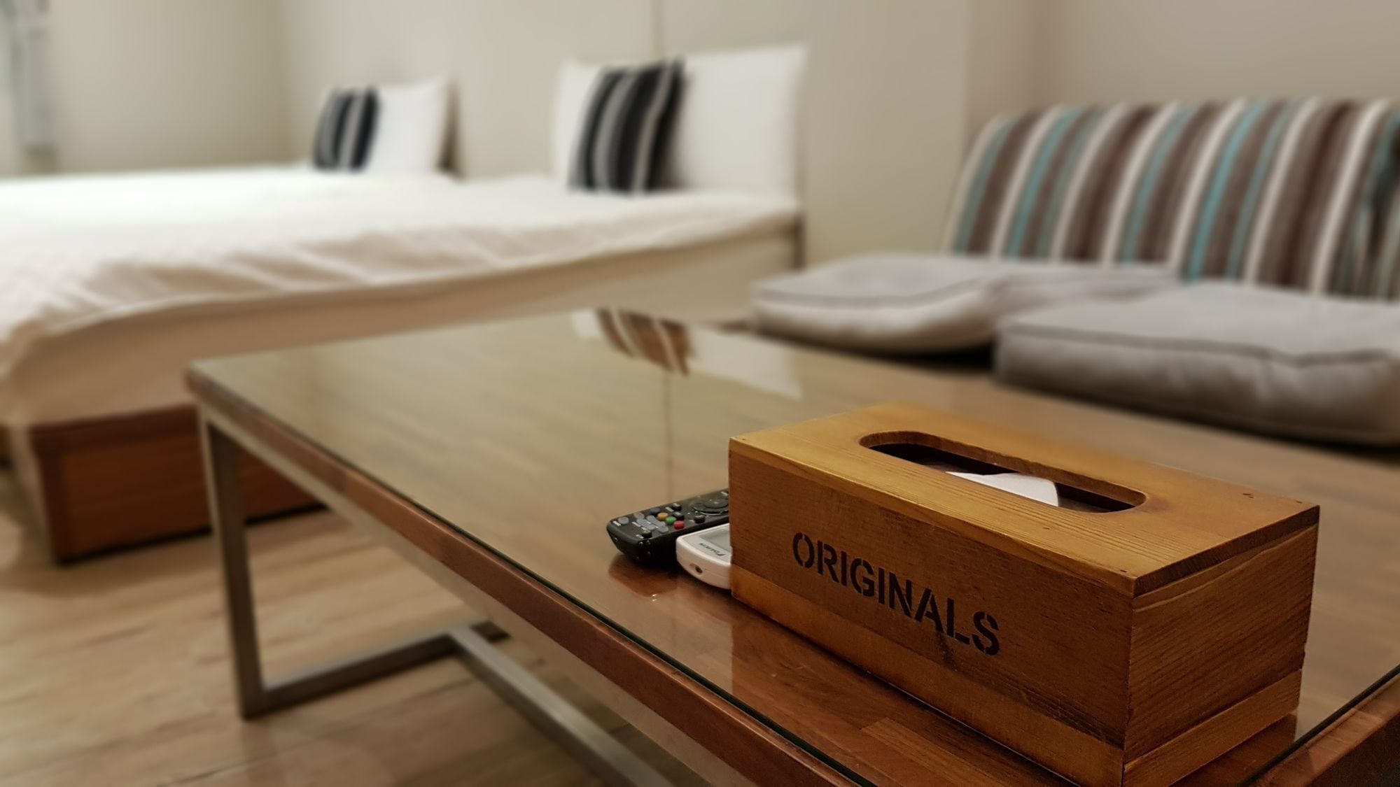 Ein Hotelzimmer mit einem Glastisch, einer hölzernen Taschentuchbox mit dem Aufdruck "ORIGINALS", einer Fernbedienung und einem gestreiften Sofa.
