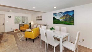 A modern living room with a dining area, featuring a large painting, a yellow armchair, and white furniture. - Snowy Pines | Walk to Snow Summit · Fireplace (Big Bear Lake)