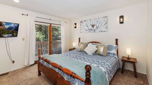 A bedroom with a wooden bed, a large window with blinds, a wall-mounted TV, and a painting. - Snowy Pines | Walk to Snow Summit · Fireplace (Big Bear Lake)