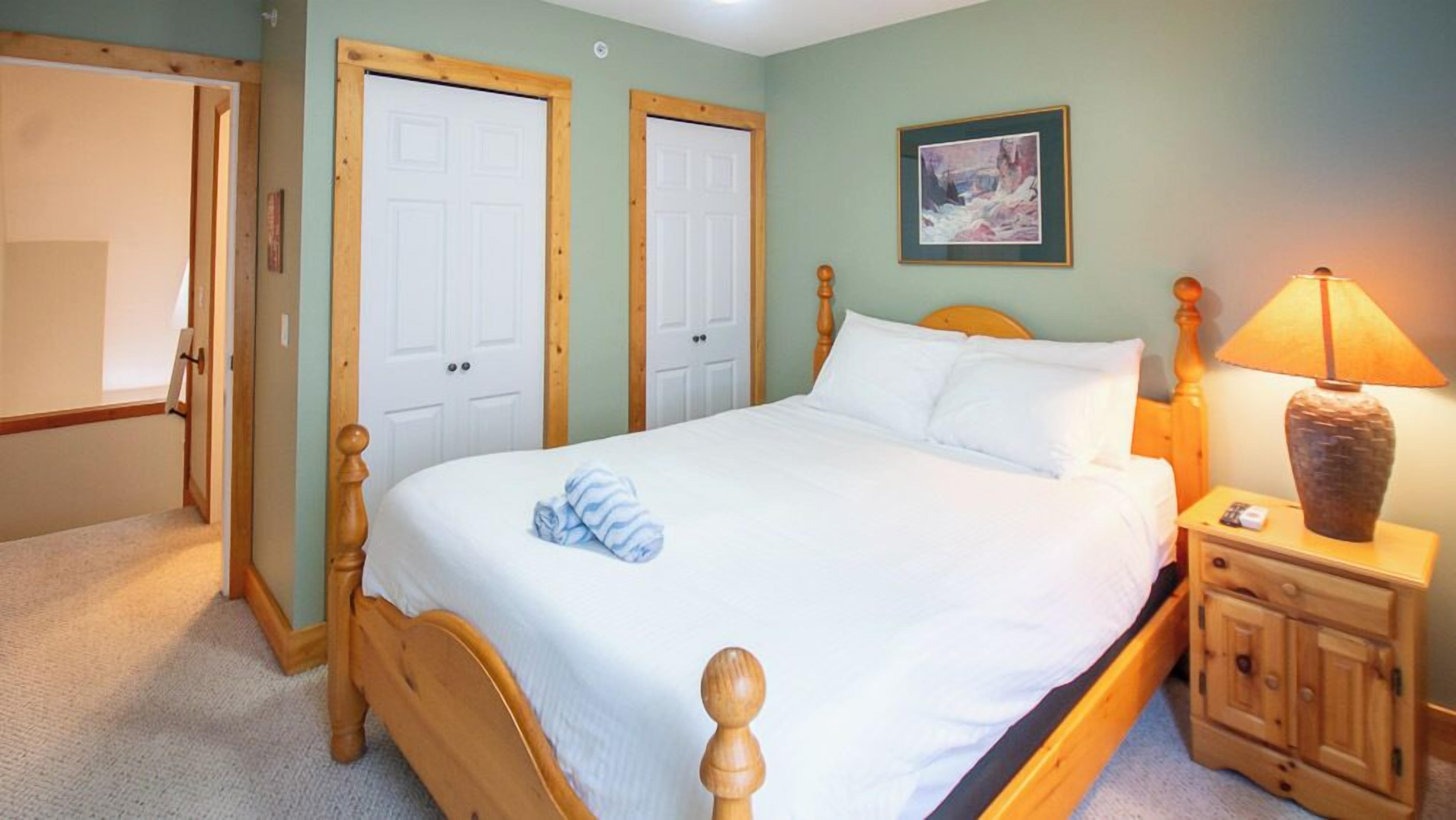 A bedroom with a wooden bed, two white doors, a painting on the wall, a nightstand with a lamp, and a small cabinet.