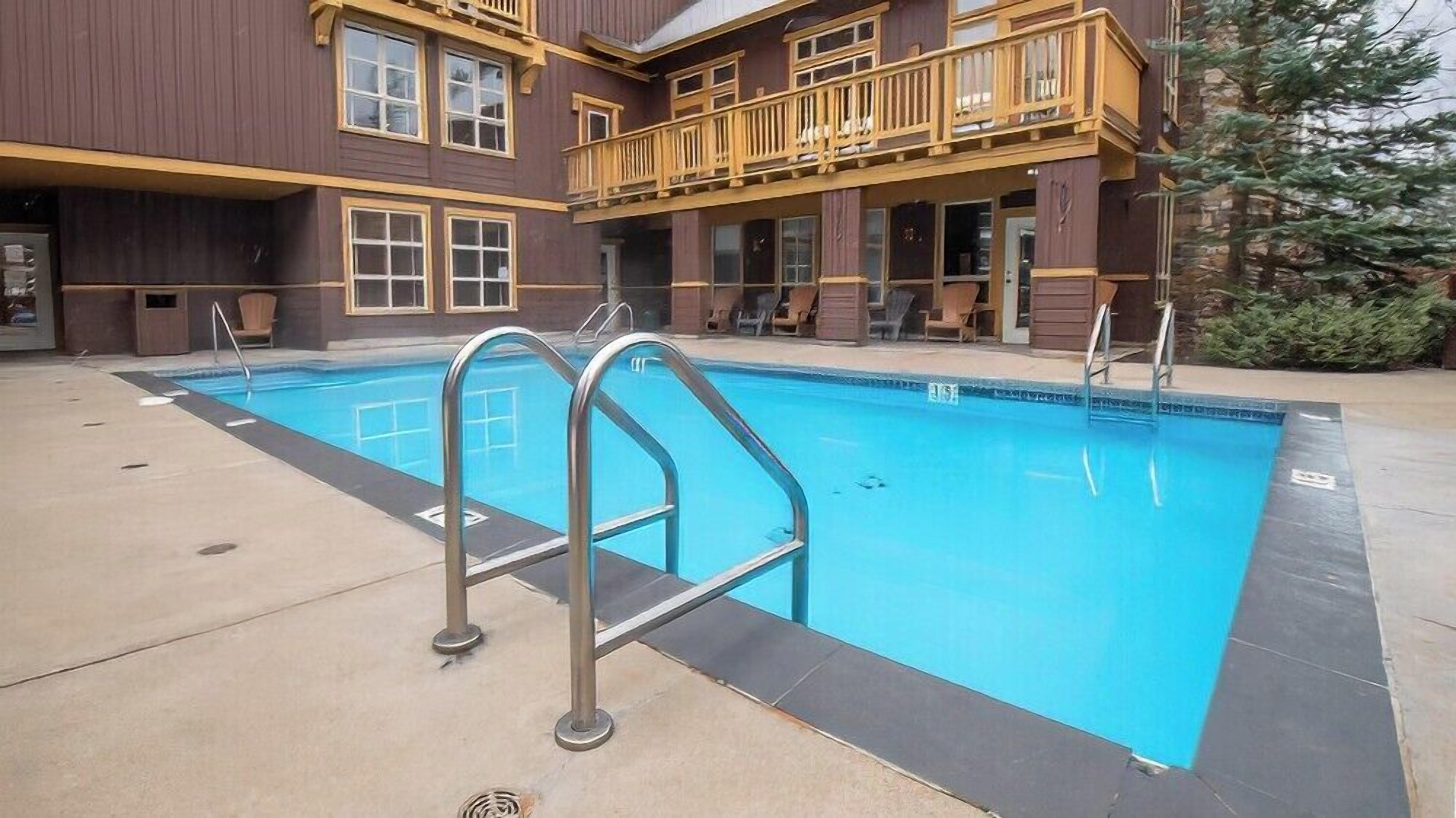 A swimming pool with two metal ladders, surrounded by a building with a wooden balcony and a stone wall.