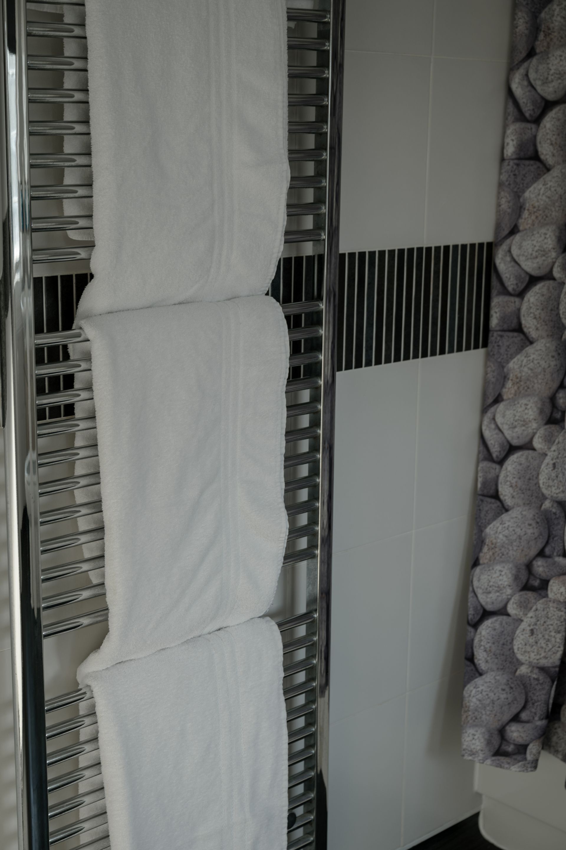 A bathroom with a towel rack holding three white towels, a tiled wall with a pebble pattern, and a shower curtain.
