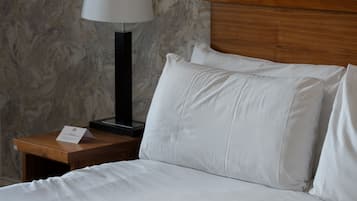 Una cama bien tendida con sábanas blancas y un cabecero de madera, una mesita de noche con lámpara, y una pared con estampado de mármol.
