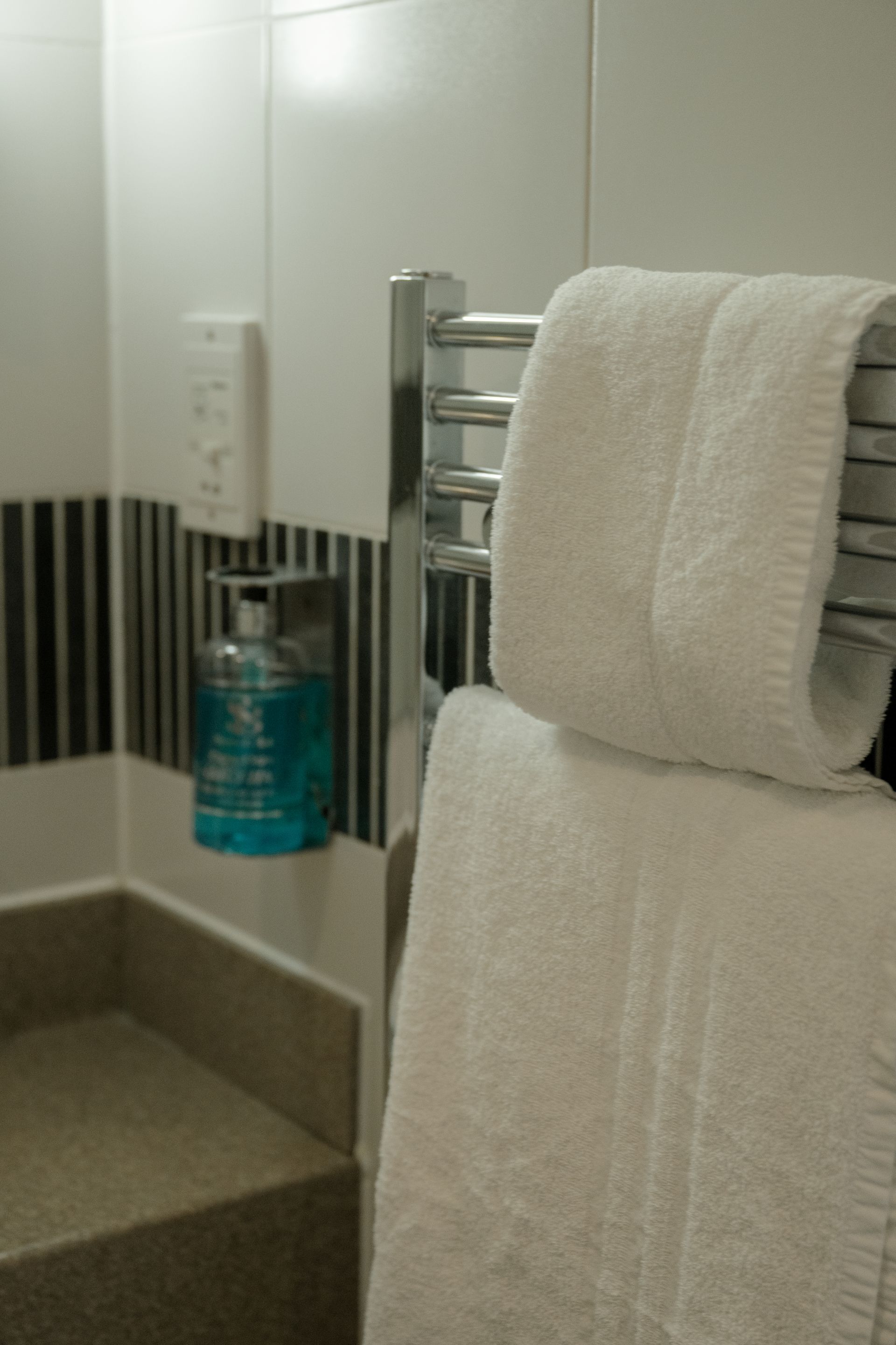 Um banheiro com um porta-toalhas com uma toalha branca, uma garrafa azul em uma prateleira e um revestimento de azulejos na parede.