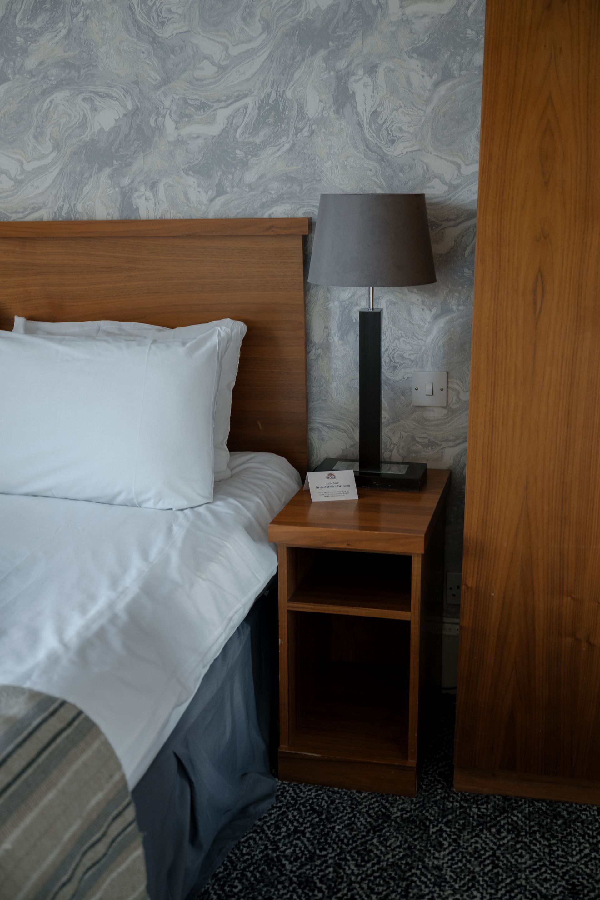 Een net opgemaakt bed met witte lakens, een houten nachtkastje met een lamp, en een wand met een marmerpatroon.