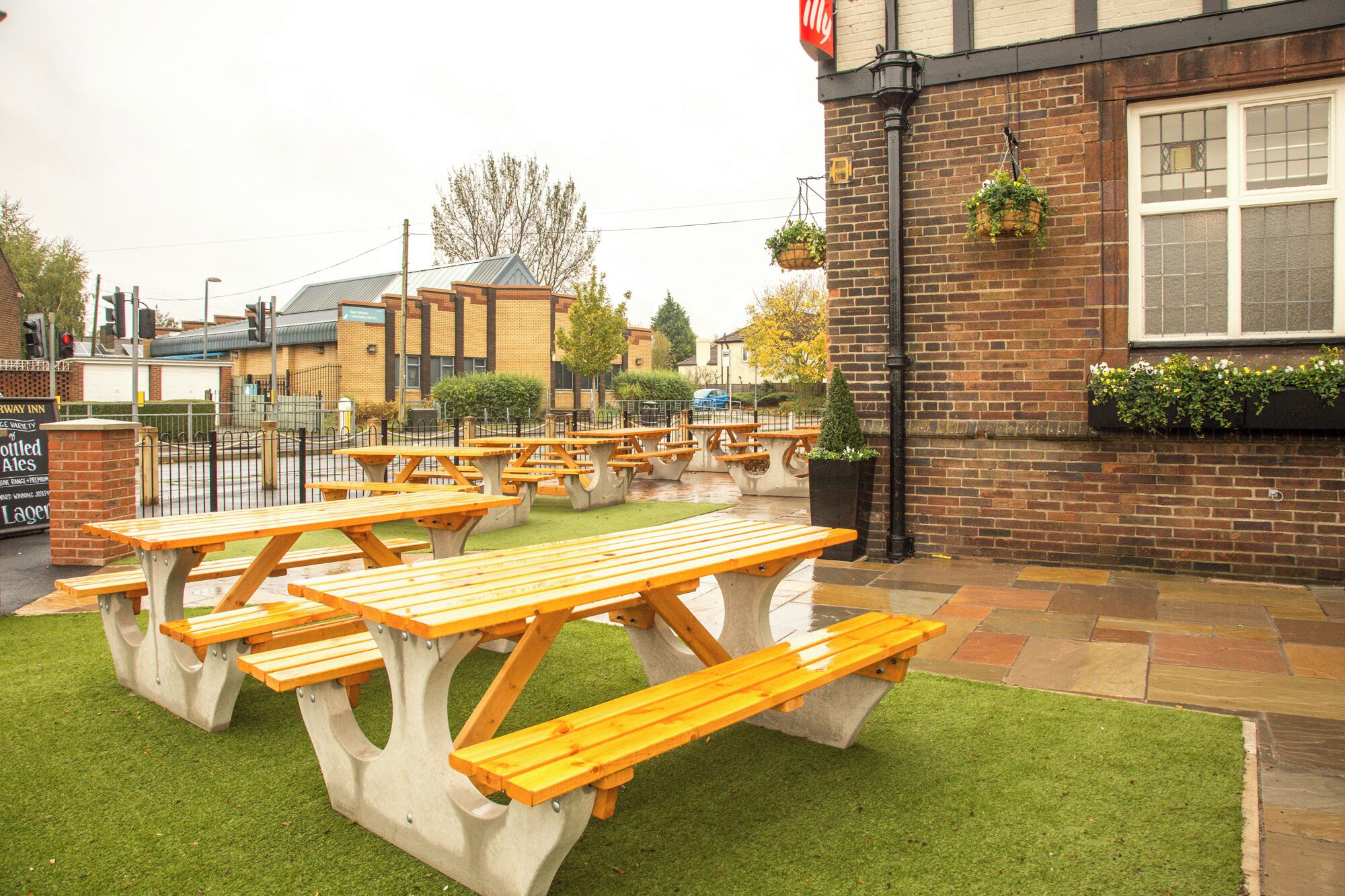 An outdoor seating area with multiple wooden picnic tables and a brick building with hanging flower baskets.