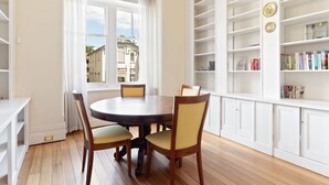 A dining area with a round table and four chairs, a bookshelf with books, and a view through a window.