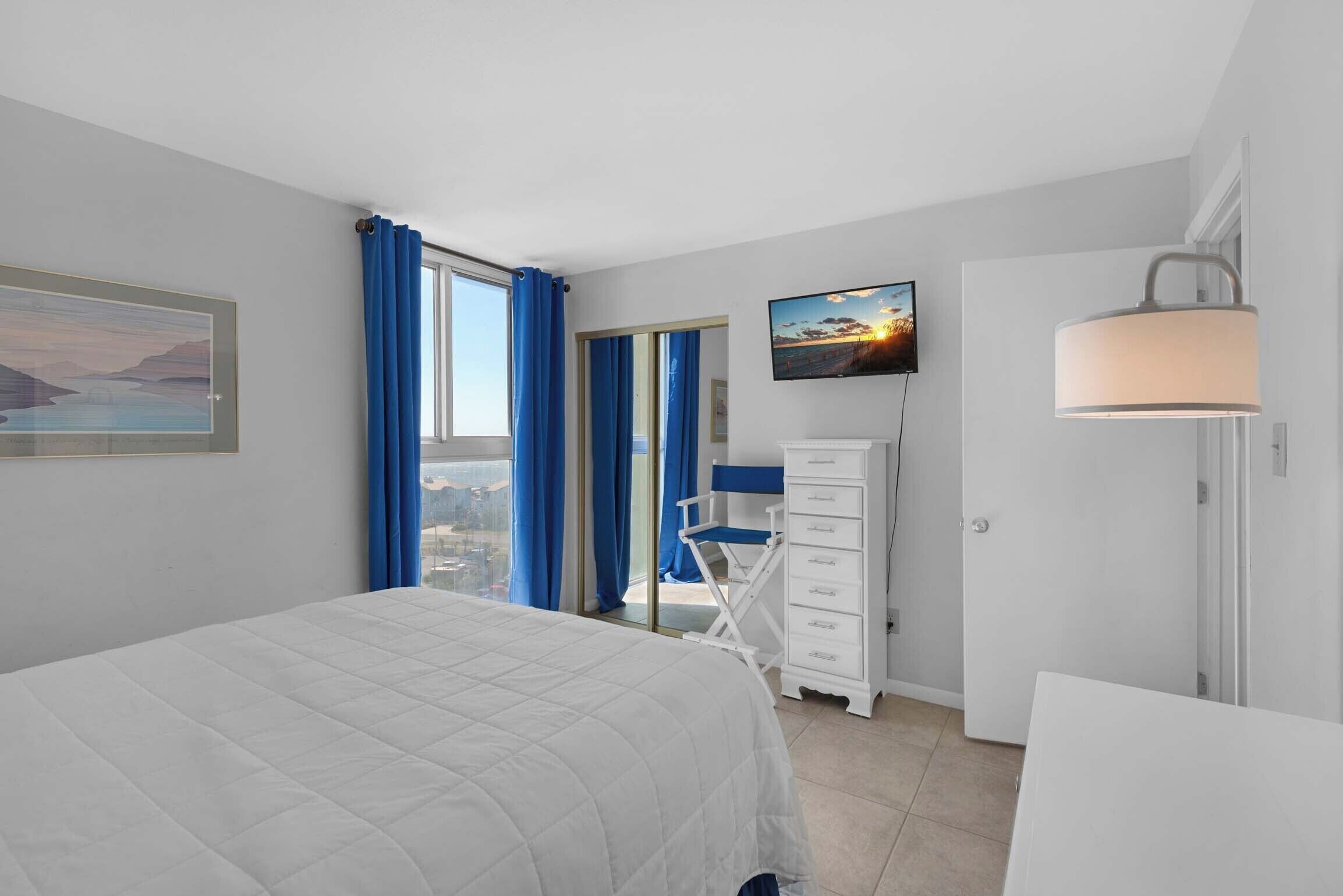 Une chambre à coucher avec un lit, un bureau, une chaise, un téléviseur et une fenêtre avec des rideaux.