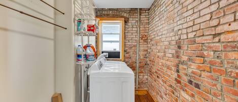 A laundry room with a white washing machine, shelves with cleaning supplies, a brick wall, and a window.