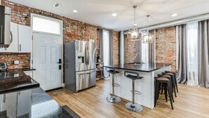 A modern kitchen with a brick wall, stainless steel appliances, a central island with bar stools, and a pendant light. - Brikctoria 1102 3 Bedroom Home (Kansas City)