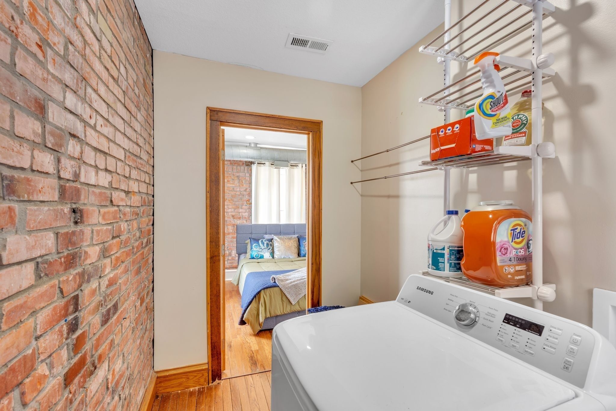 A laundry area with a washing machine, shelves with cleaning products, and a brick wall.