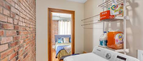 A laundry area with a washing machine, shelves with cleaning products, and a brick wall.