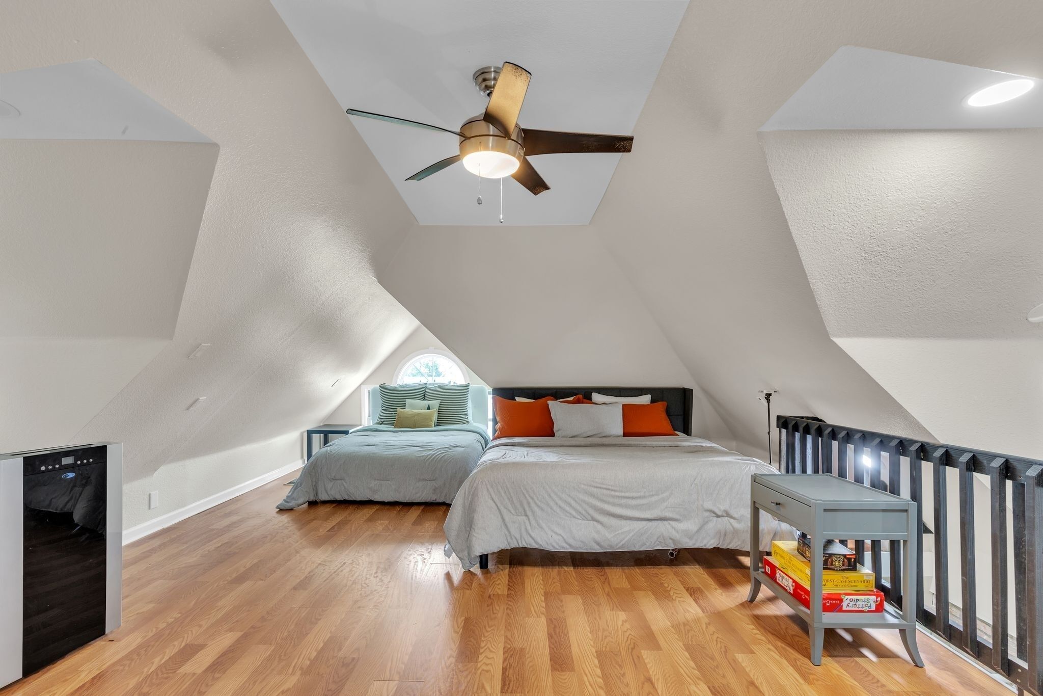 A bedroom with a sloped ceiling, a ceiling fan, a bed with orange and white bedding, a bedside table with books, and a window with a view.