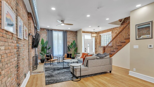 A spacious living room with a brick wall, a wooden staircase, and a grey sofa. - Brikctoria 1102 3 Bedroom Home (Kansas City)