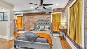 A bedroom with a brick wall, a tufted headboard, a bed with an orange throw, and a desk with a lamp. - Brikctoria 1102 3 Bedroom Home (Kansas City)