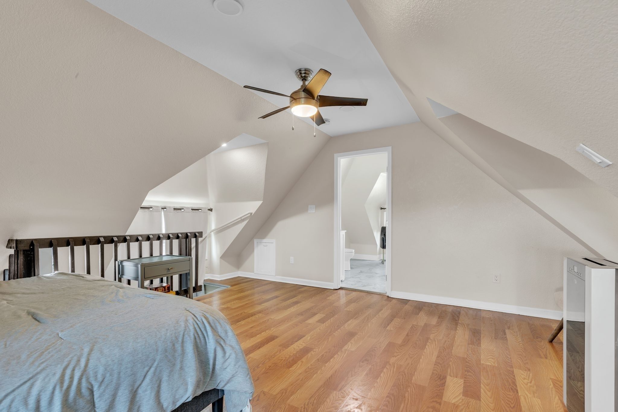 A bedroom with a wooden floor, a bed with a grey blanket, a ceiling fan, and a white door leading to a bathroom.