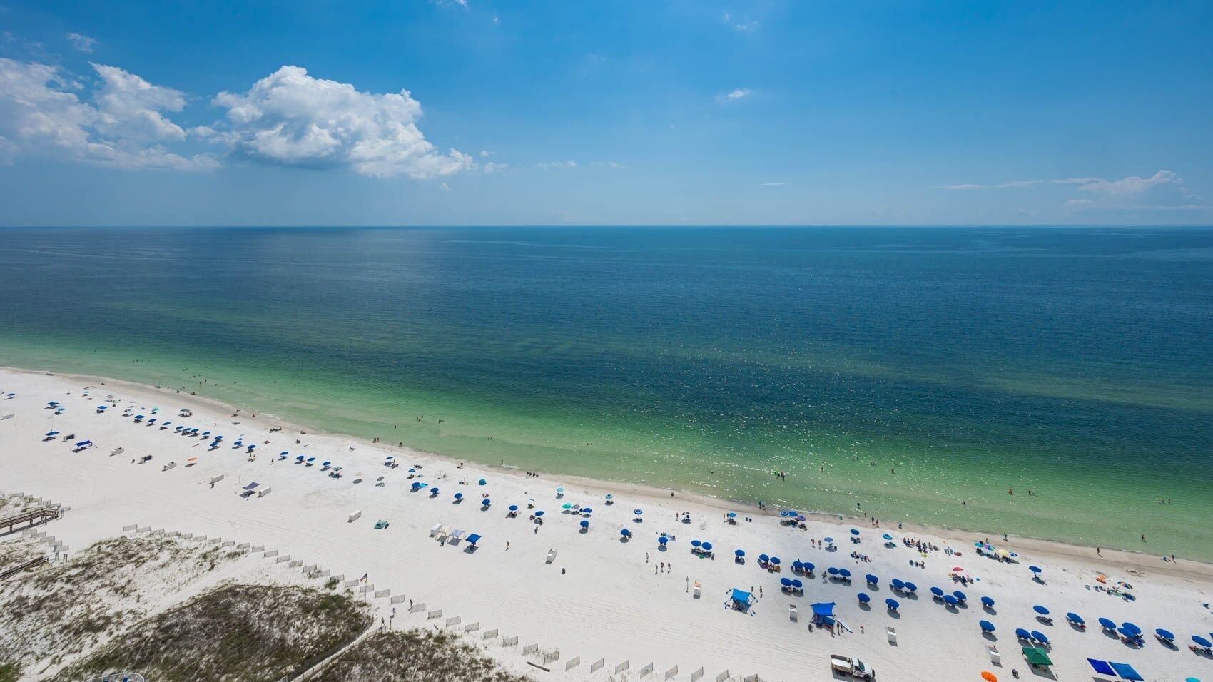 A beach with white sand, clear blue water, and numerous blue umbrellas.
