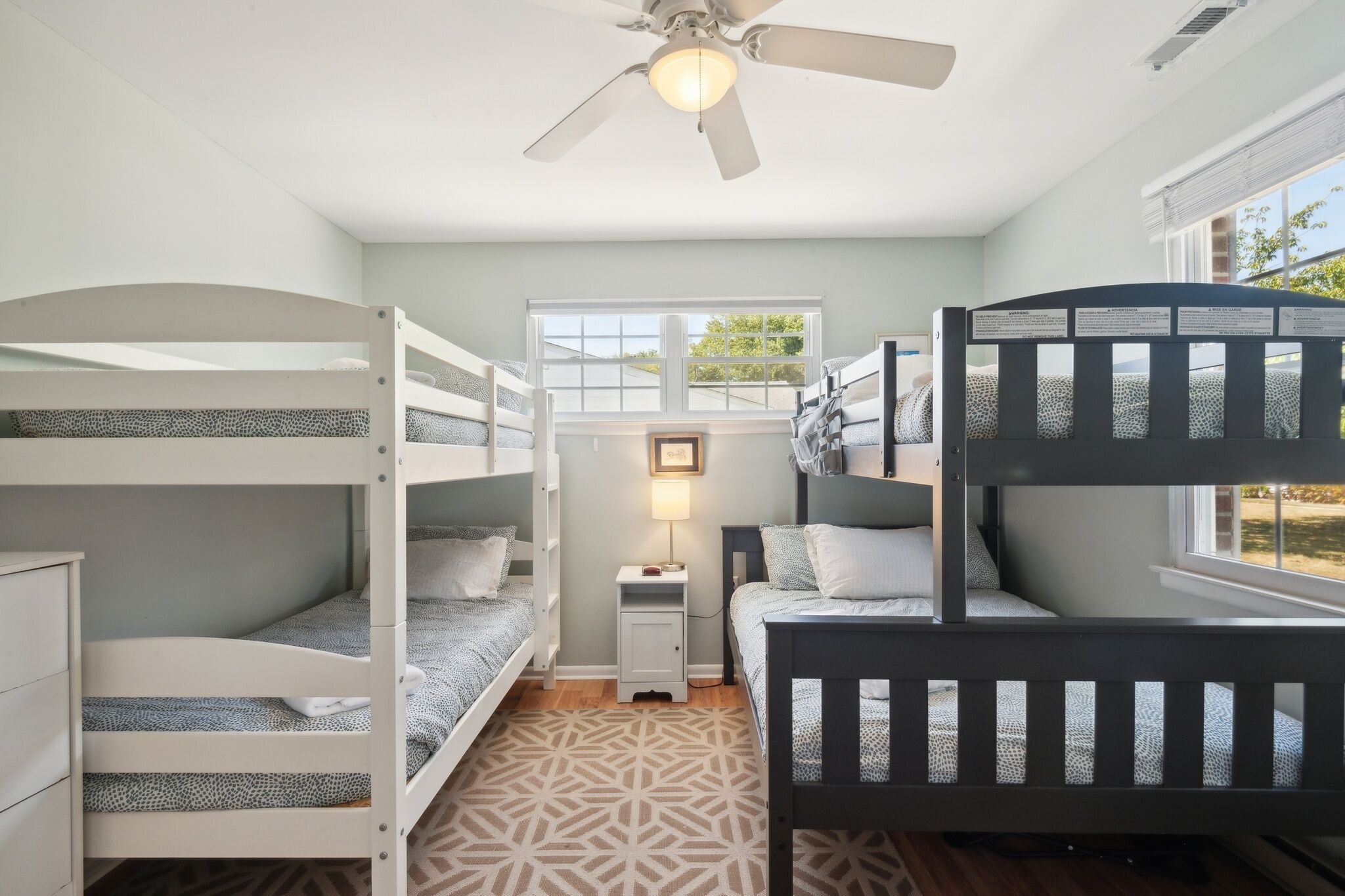 A bunk bed room with a ceiling fan, a small bedside table with a lamp, and a window with a view of greenery.
