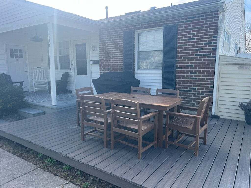 A wooden deck with a table and chairs, a barbecue grill, and a brick house with a porch.