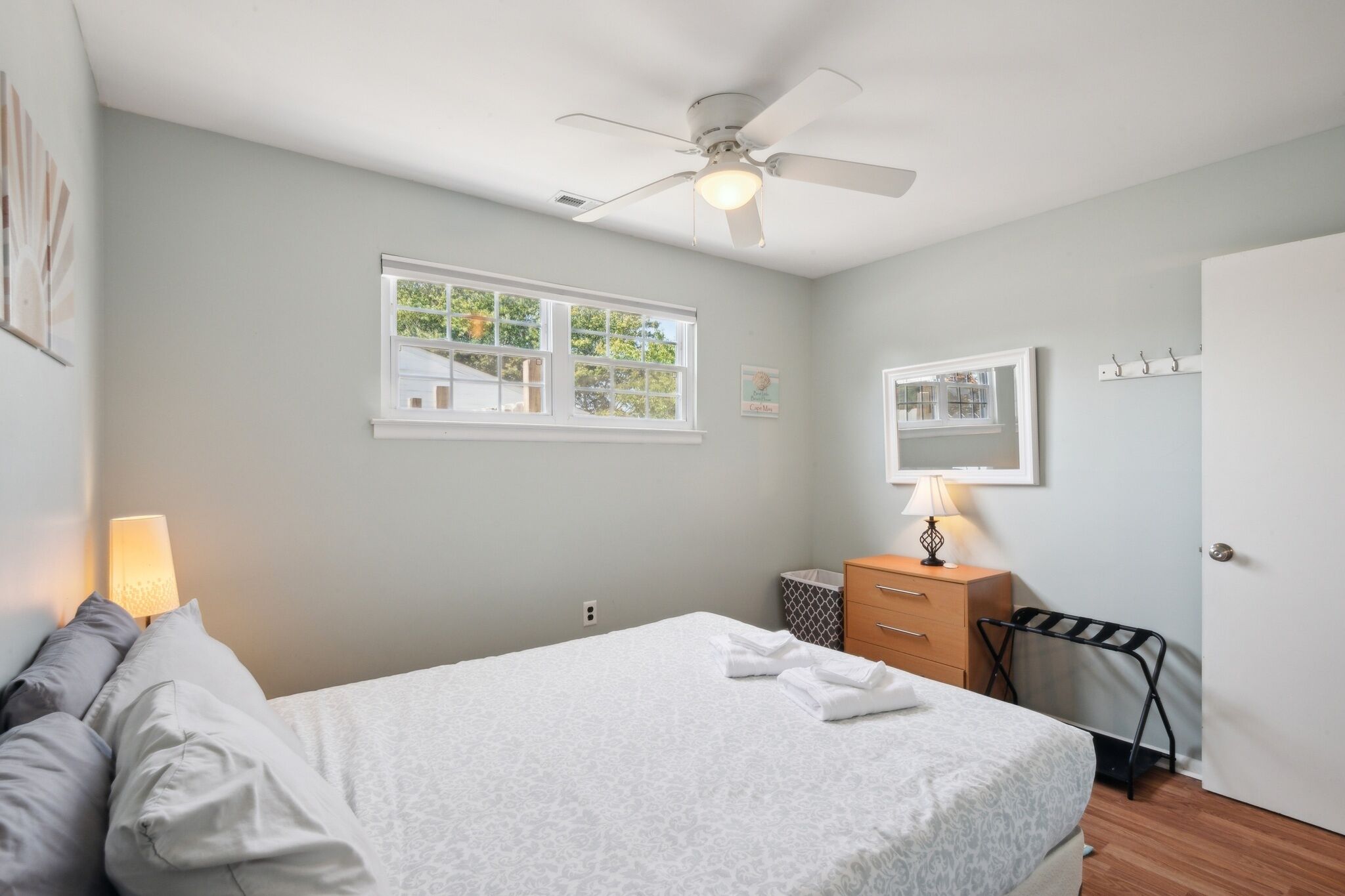 A bedroom with a bed, a ceiling fan, a window, a bedside lamp, a dresser, and a door.