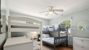 Un dormitorio infantil con literas, un ventilador de techo, una mesita de noche con lámpara y un aparador.