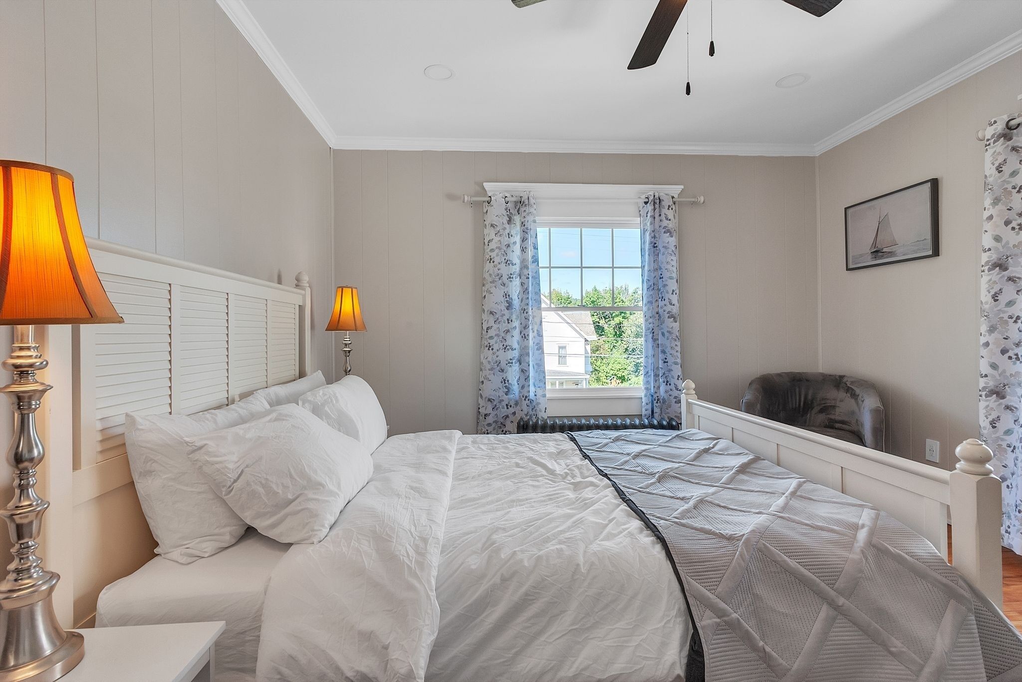 Une chambre à coucher avec un lit, deux lampes de chevet, une fenêtre avec des rideaux, une chaise et un tableau encadré au mur.