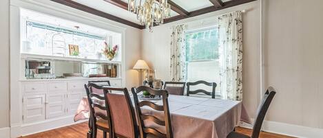 A dining room with a wooden table, chairs, and a chandelier. There is a window with curtains and a view of greenery outside.