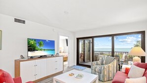 A modern living room with a flat-screen TV, a sofa, a coffee table, and a balcony with a view of the sea.