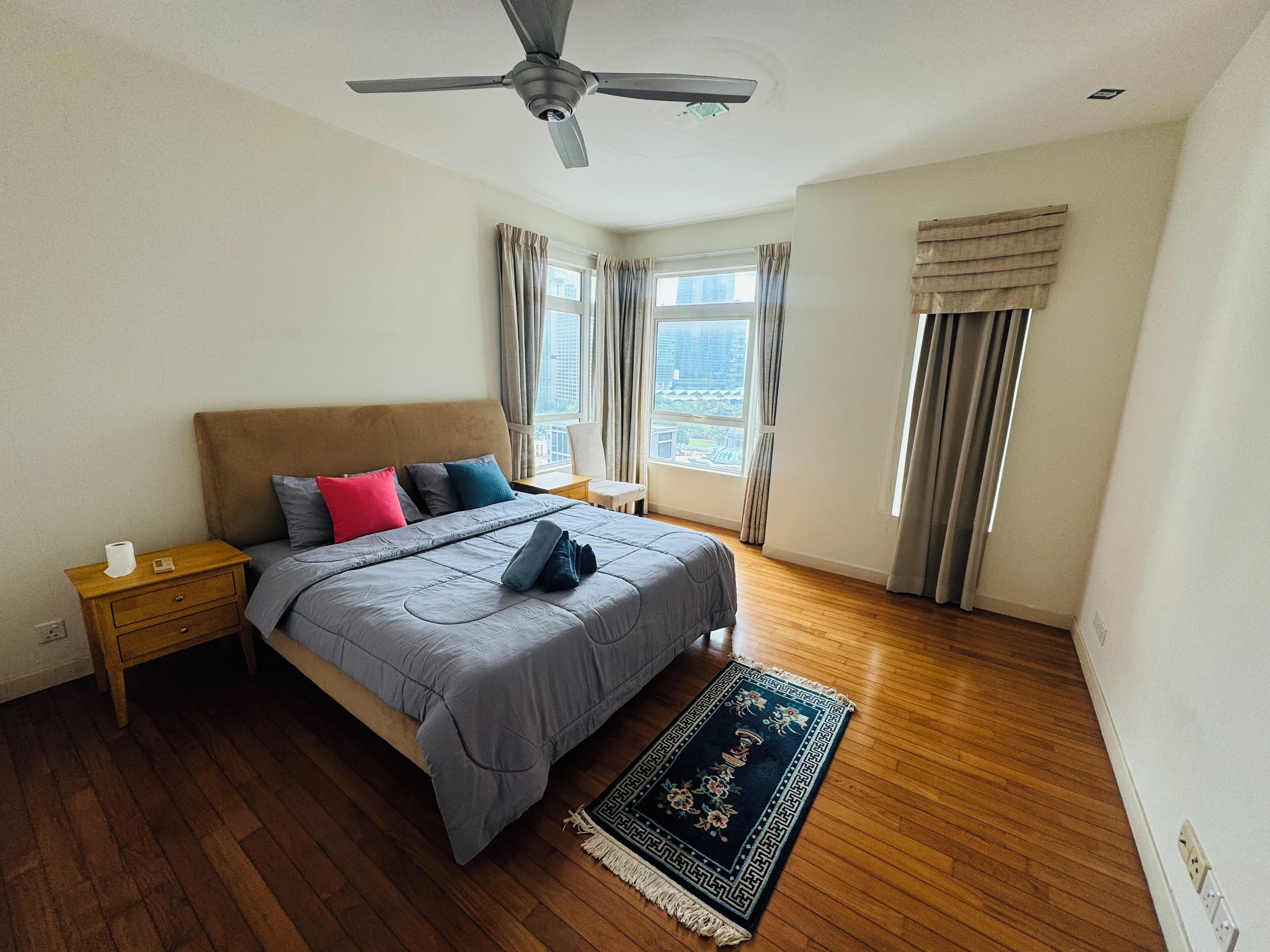 Un dormitorio con cama, piso de madera, ventilador de techo, ventana con cortinas y una mesita de noche con lámpara.