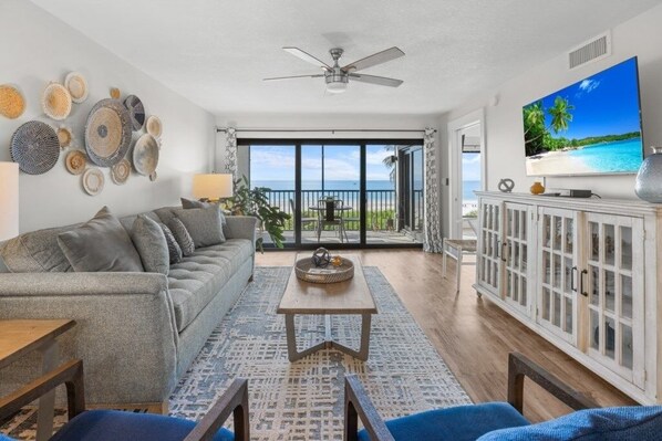 A living room with a sofa, coffee table, and TV, a balcony with a view of the ocean, and a ceiling fan.