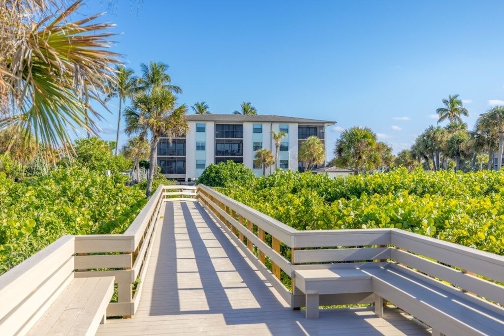 A wooden boardwalk leading to a building with multiple floors, surrounded by lush greenery and palm trees.
