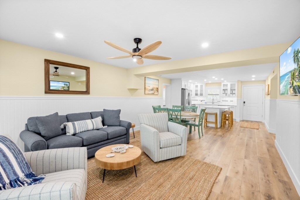 A spacious living room with a grey sofa, a striped armchair, and a round coffee table. A ceiling fan is visible, and the room has wooden flooring.