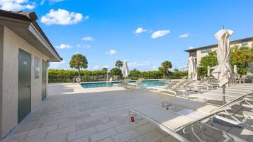 A swimming pool area with lounge chairs and umbrellas, a tiled deck, and a building with a door and a sign.