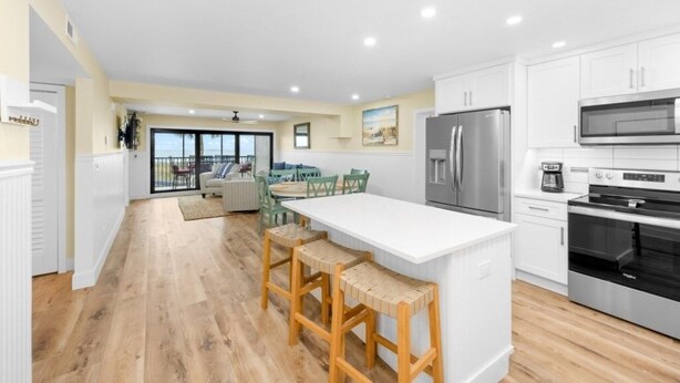 A modern kitchen with white cabinets, a stainless steel refrigerator, and a built-in oven. There is a dining area with a table and chairs, and a large window providing a view of the outdoors. - Tarpon Beach 302 3 Bedroom Condo (Sanibel)