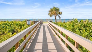 A wooden boardwalk leading to a beach with a palm tree and lush greenery. - Tarpon Beach 302 3 Bedroom Condo (Sanibel)