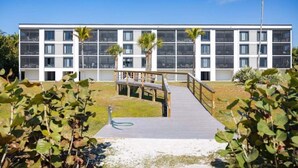 A modern building with a wooden deck leading to a gated entrance, surrounded by greenery. - Pelicans Roost 105 2 Bedroom Condo (Sanibel)
