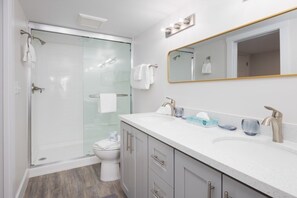 Un baño moderno con cabina de ducha de vidrio, inodoro blanco, tocador con lavamanos y espejo, y una pequeña encimera con artículos de aseo.