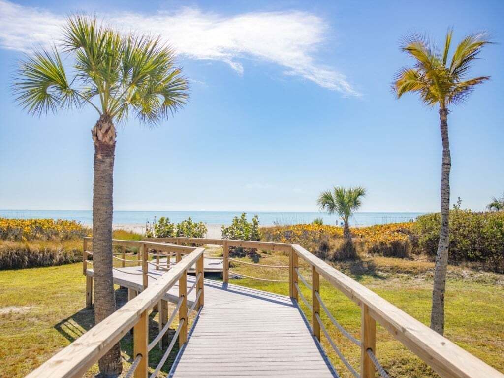 Een houten looppad dat naar een strand leidt, met palmbomen en een heldere lucht.