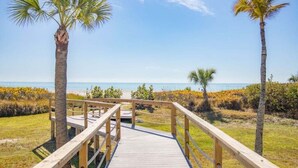 A wooden boardwalk leading to a beach with palm trees and a clear sky. - Pelicans Roost 105 2 Bedroom Condo (Sanibel)