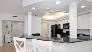 A modern kitchen with white cabinetry, a black refrigerator, and a central island with two white chairs. - Captivated By The Sea 2 Bedroom Condo (Captiva)