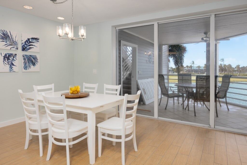 Un coin repas avec une table et des chaises blanches, un lustre, et une vue sur une terrasse extérieure accessible par une porte vitrée, où se trouvent des chaises et une table.