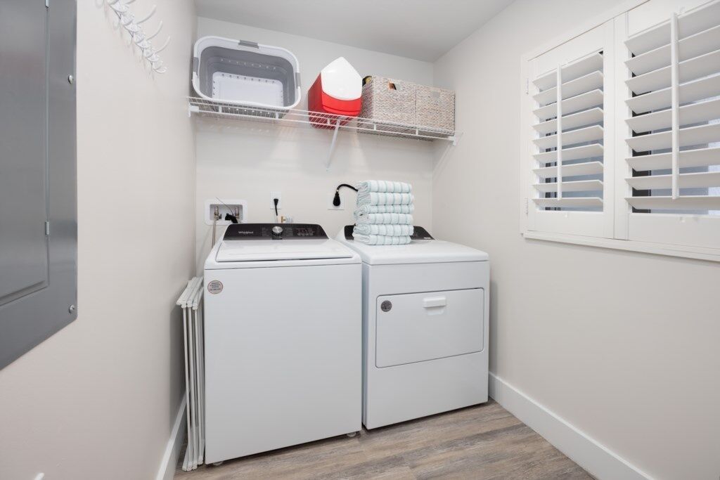 A laundry room with a front-loading washer and dryer, a shelf with baskets, and a window with blinds.
