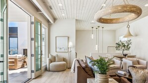 A modern living room with a large sofa, a coffee table, and a dining area. There is a large window providing a view of the outdoors. - Rosemary 7 Bedroom Home (St. George)