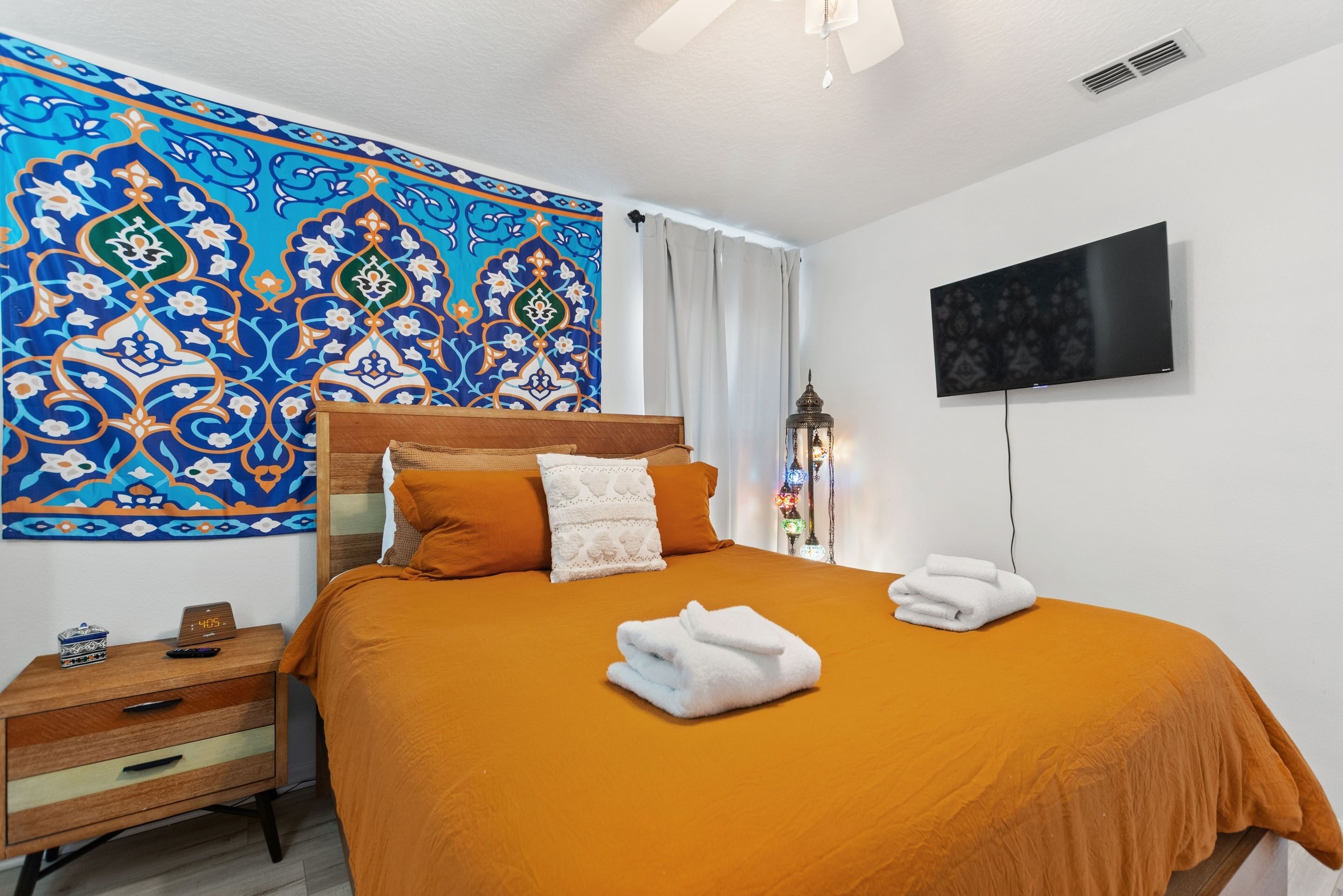 A hotel room with a large bed, orange bedding, a TV on the wall, a nightstand with a clock, and a decorative wall tapestry.