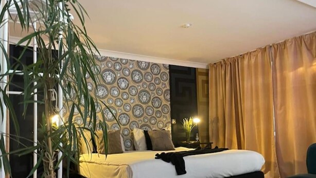 A modern bedroom with a bed, bedside tables, a potted plant, and a patterned wallpaper. - The Exclusive Jacuzzi Apartment (Birmingham)