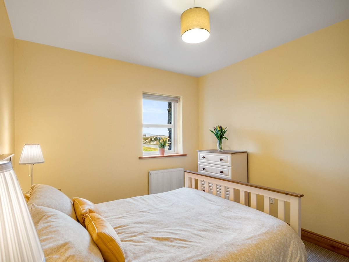 A bedroom with a bed, bedside lamp, dresser, window, and a view of the outdoors.